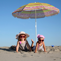 Zwei Mädchen mit Sonnenbrille und Sonnenhut liegen am Strand unter einem Sonnenschirm.